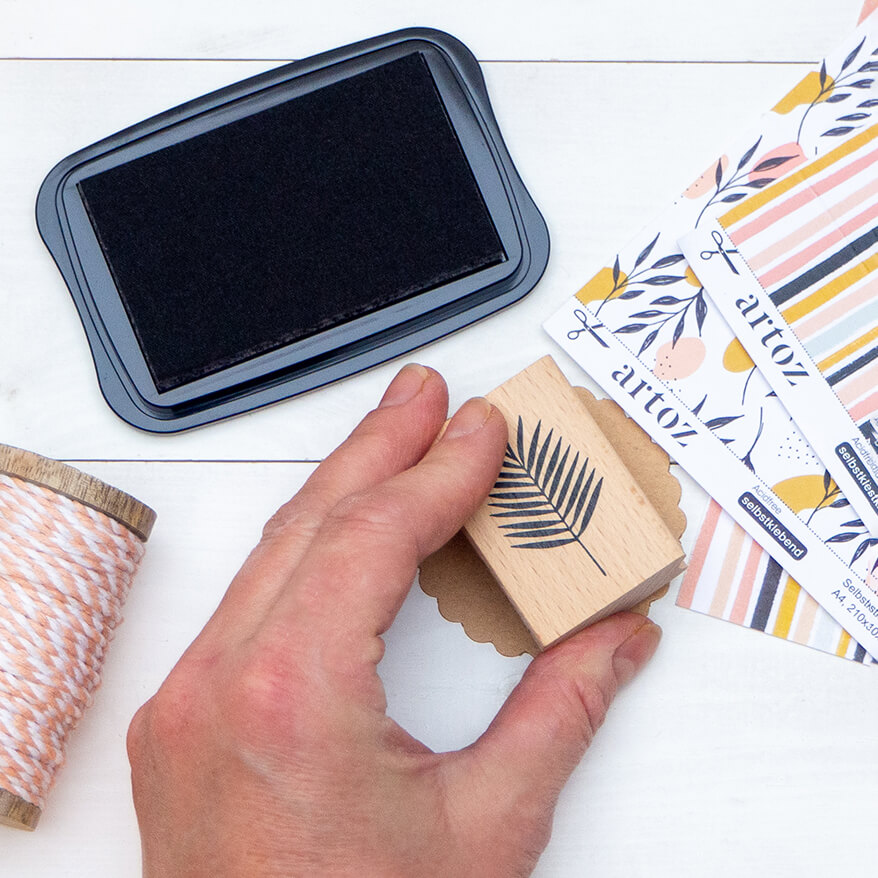 Eine Hand stempelt mit einem Stempel der Marke Artoz ein fiedriges Blatt auf ein rundes Etikett aus Kraftpapier.