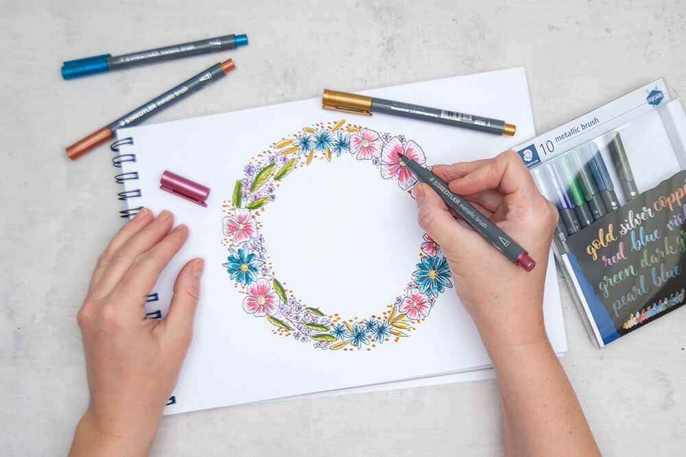 Jemand hebt mit den metallic brush Markern von Staedtler einzelne Highlights in einem gemalten Blumenkranz hervor. Foto: Johanna Rundel