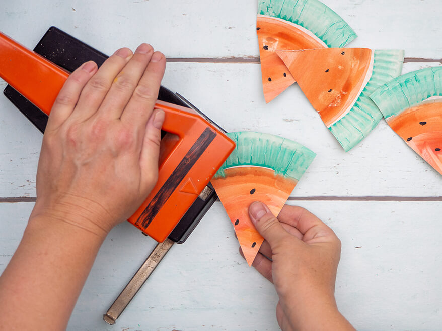 Jemand stanzt mit einem orangefarbenen Locher Löcher in die Ecken eines Dreiecks. Dieses besteht aus einem Pappteller, der wie eine Wassermelone angemalt ist und als Dreieck ausgeschnitten wurde. Der Untergrund ist hell. Foto: Johanna Rundel 