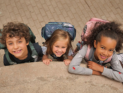 Ein Junge und zwei Mädchen im Grundschulalter stehen mit ihren Schulranzen von School-Mood vor einer beigen Mauer und blicken lachend nach oben in die Kamera. Das Mädchen in der Mitte hält sich an der Mauer fest, das Mädchen rechts stützt sich mit den Armen darauf ab.