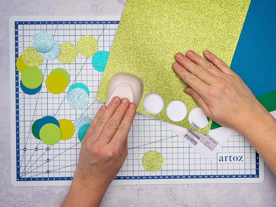 Jemand stanzt mit einem Kreisstanzer Kreise aus verschiedenen Papieren in Grün- und Blautönen. Der Untergrund ist eine helle, faltbare Schneidematte mit Linien von Artoz. Foto: Johanna Rundel