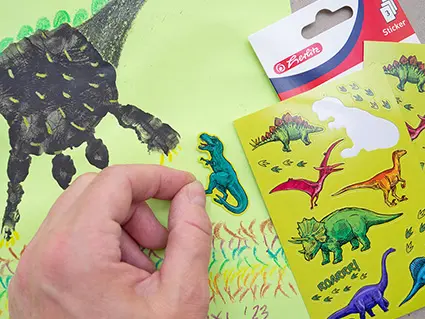 Jemand klebt einen Dinosaurier-Sticker von einem Stickerbogen von Herlitz auf ein selbstgestaltetes Dino-Bild aus Handabdrücken. Foto: Johanna Rundel