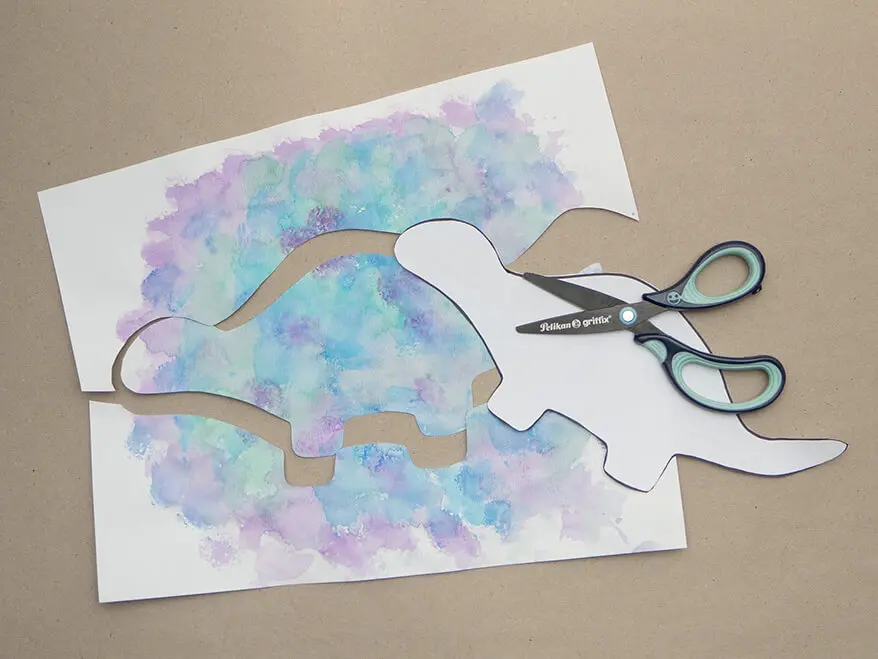 Ein Dinosaurier-Umriss wird aus einem, mit Wasserfarbe von Pelikan betupftem, Papier ausgeschnitten. Foto: Johanna Rundel