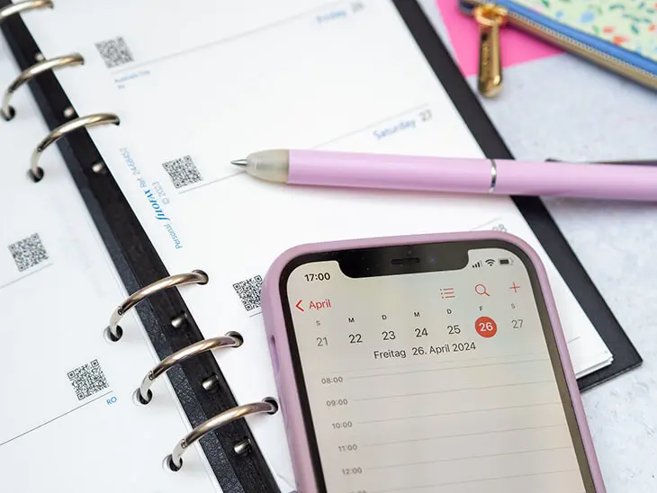 Auf einem Planer von Filofax liegt ein lila Stift und ein Smartphone, bei dem der Kalender geöffnet ist. Foto: Johanna Rundel