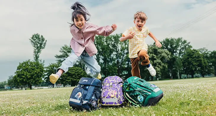Ein Mädchen und eine Junge im Grundschulalter springen auf einer grünen Wiese beherzt über ihre Schulranzen von ergobag.