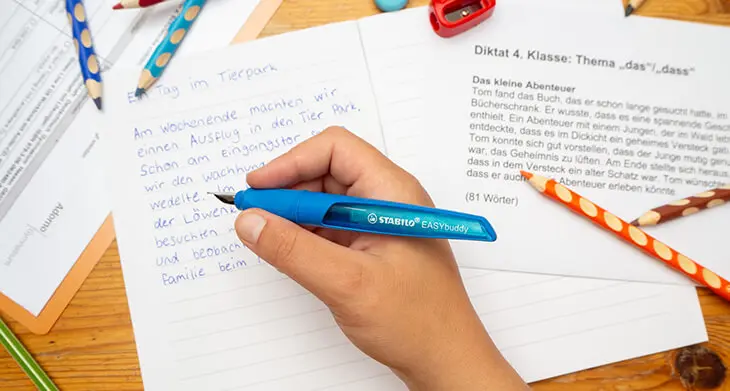 Jemand schreibt mit einem blauen EASYbuddy Füllfederhalter von STABILO ein Diktat in ein Heft. Verschiedene Papiere und weiteres Schulmaterial sind drumherum verteilt. Foto: Johanna Rundel