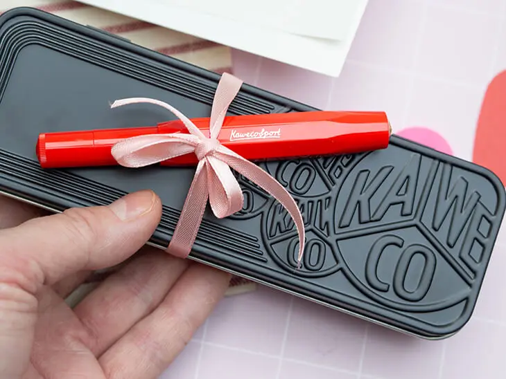 Jemand hält eine schwarze Metallbox von Kaweco in der Hand. Darauf ist mit einer rosa Satinschleife ein roter Füller befestigt. Foto: Johanna Rundel