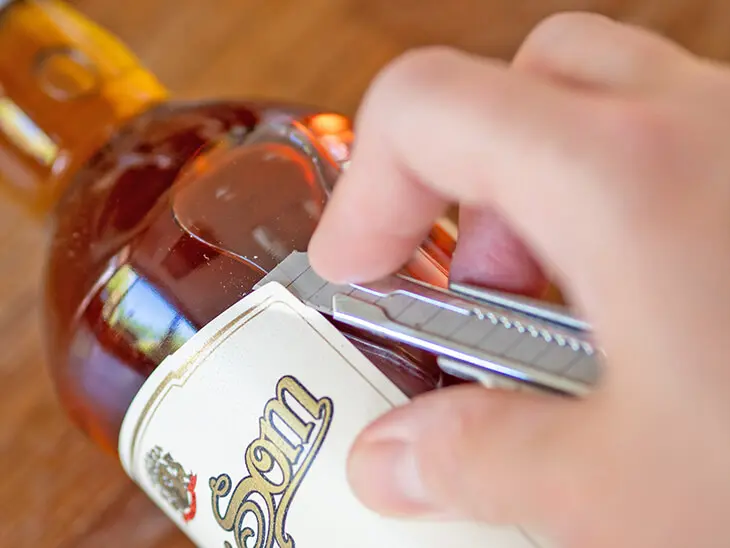 Jemand löst vorsichtig  mit einem Cuttermesser das Etikett einer Spirituosenflasche ab. Foto: Johanna Rundel