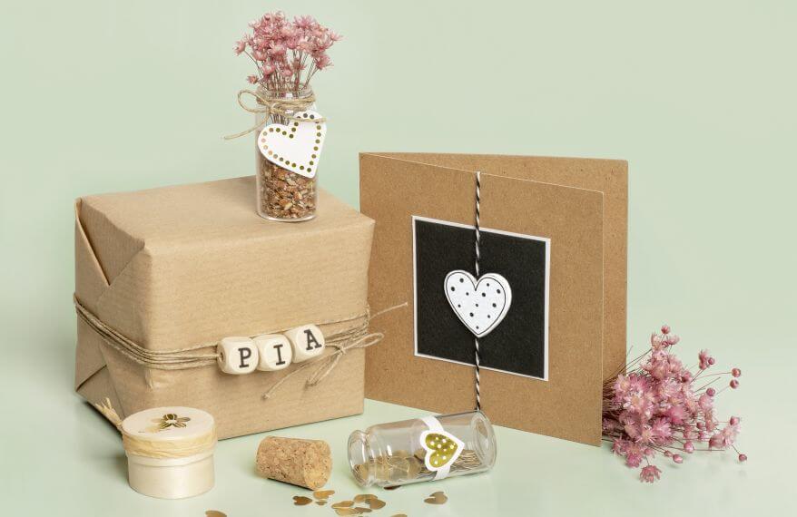 Vor grünem Hintergrund steht ein in Kraftpapier eingepacktes Geschenk. Es ist umwickelt mit einer Kordel, auf dem Würfel mit dem Namen "Pia" zu aufgefädelt sind. Darauf steht ein kleines, dekoriertes Einweckglas mit Trockenblumen. Daneben ist eine selbstgebastelte Grußkarte mit Herz zu sehen, drumherum weitere Dekoration. Das Ensemble wurde mit Rubbelstickern der Marke Heyda gestaltet.