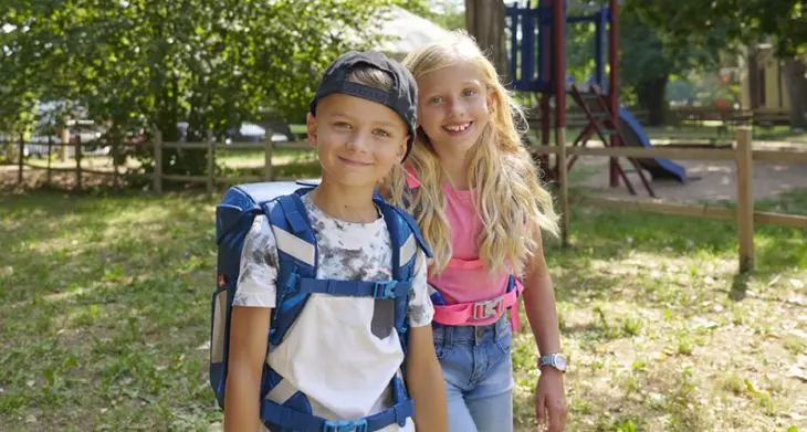 Zwei Kinder mit Schulranzen von Scout auf dem Rücken stehen auf einem Spielplatz und lächeln.