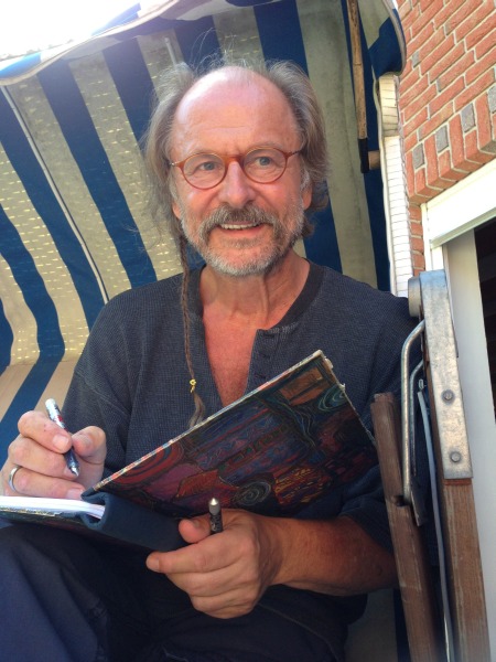 Klaus-Peter Wolf sitzt zuhause in seinem Strandkorb und hält ein Notizbuch in der Hand, in welches er gerade schreibt.