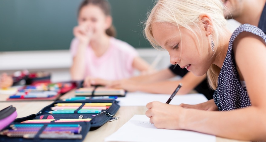 Ein Mädchen sitzt in der Schule am Tisch uns schreibt etwas. Vor ihr liegt ein aufgeschlagenes Federmäppchen . Im Hintergrund ist ein anders Mädchen zu erkennen.