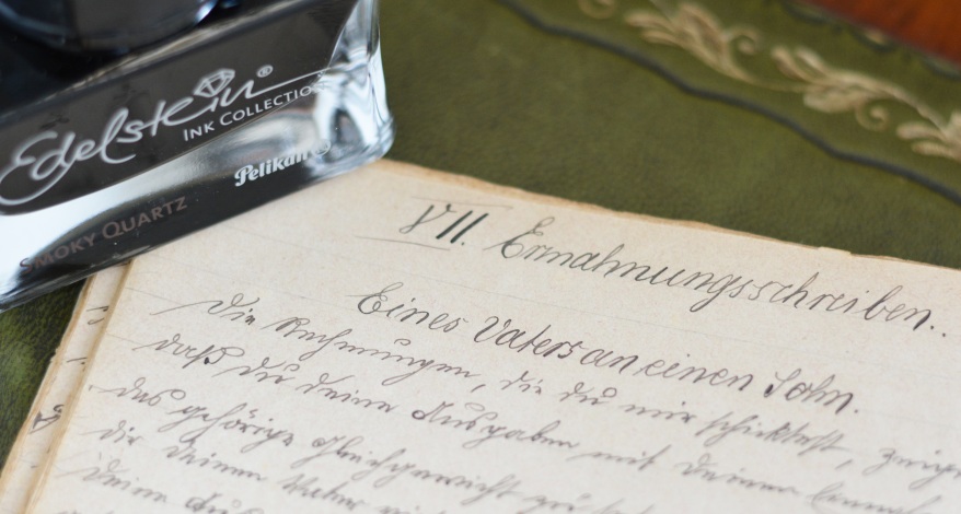 Ein altes Schriftstück mit dem Titel "Ermahnungschreiben" ist zu sehen. Der Brief wurde mit schwarzer Tinte auf liniertem Papier verfasst. Das Papier ligt auf einer grünen Tischdecke. In der oberen Bildecke ist eine schwarze Tinte im Glas von Pelikan zu sehen. Sie gehört zu der Edelstein Ink Collection.
