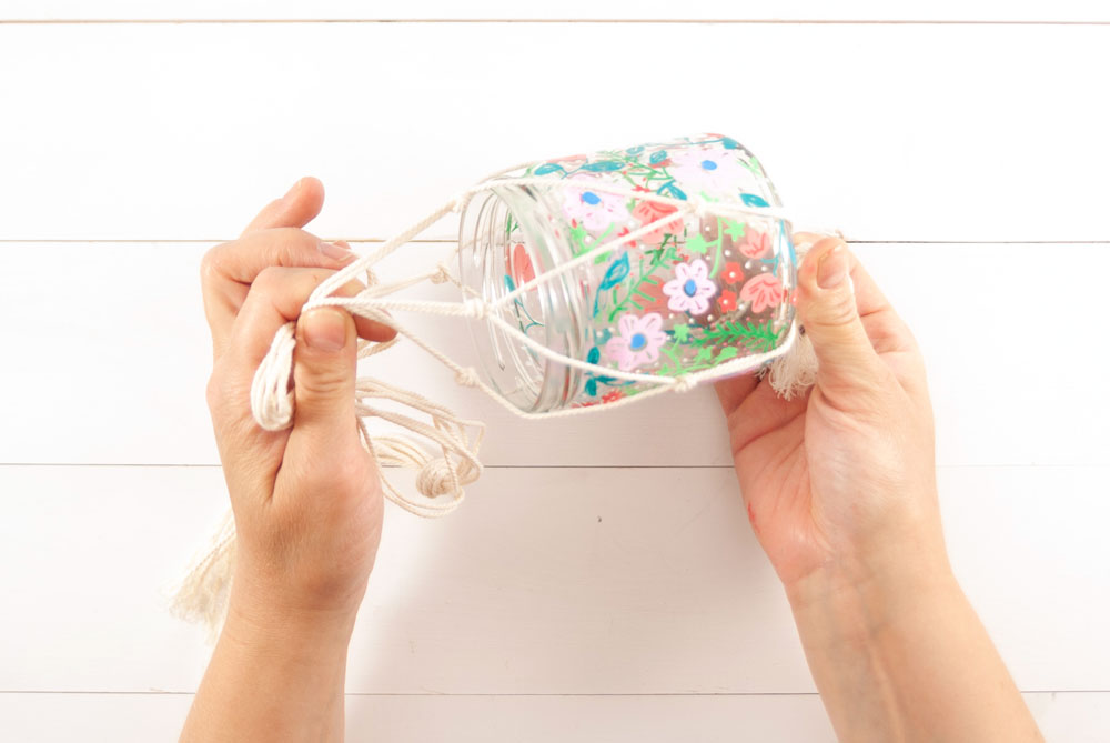 Ein bunt bemaltes Glas wird in einer Makramee-Ampel platziert.