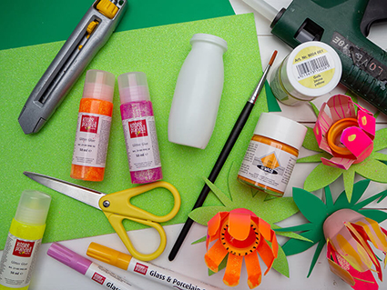 Auf einem hellen Untergrund liegt Material von Knorr Prandell und Heyda verteilt, dass man zu Herstellung einer Upcycling-Blumenlichterkette braucht: kleines Plastikfläschchen, Acrylfarben, Acrylmarker, Glitzerkleber, Moosgummi, Lichterkette, Pinsel, Schere, Cutter und eine Heißklebepistole. Man sieht auch einen Teil der fertigen Lichterkette ins Bild ragen. Foto: Johanna Rundel