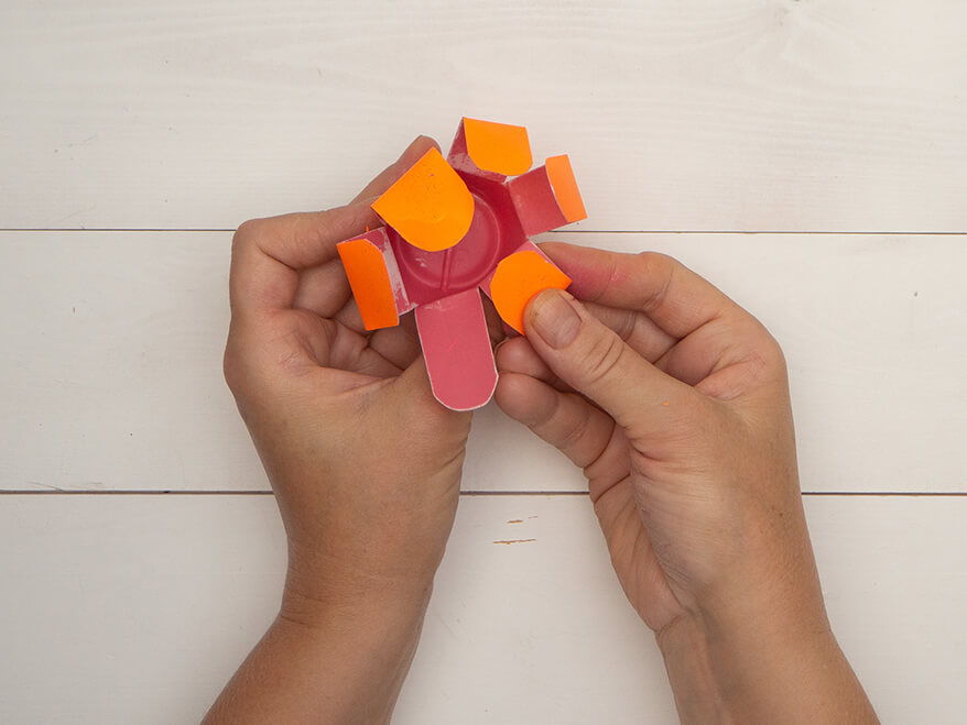 Jemand knickt die Streifen einer in Streifen geschnittenen, halben, orange bemalten, kleinen Plastikflasche erst einmal nach außen, dann nach innen um. Eine Blume entsteht. Der Untergrund ist hell. Foto: Johanna Rundel