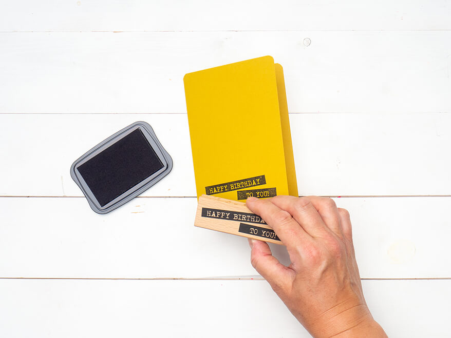 Jemand stempelt, mit Hilfe eines Stempels und eines schwarzen Stempelkissens von Artoz, die Worte "Happy Birthday'" auf eine gelbe Blanko Klappkarte. Der Untergrund ist weiß. Foto: Johanna Rundel 