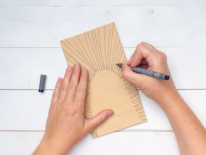 Jemand zeichnet mit einem dünnen, schwarzen Stift, strahlenförmig Striche auf eine braune Papiertüte von Artoz. Der Untergrund ist weiß. Foto: Johanna Rundel 