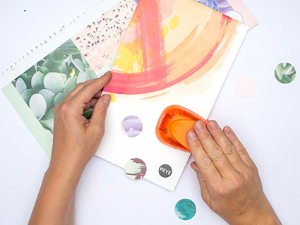 Jemand stanzt mit einer orangefarbenen Stanze Kreise aus alten Kalenderblättern von Athesia aus. Der Untergrund ist weiß. Foto: Johanna Rundel