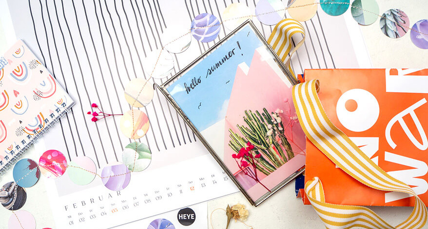 Auf einem hellen Untergrund liegen verstreut verschiedene, selbstgemachte Basteleien aus alten Kalenderblättern von Athesia. Eine DIY-Papiertüte, eine Postkarte im silbernen Bilderrahmen, eine Girlande aus aneinander genähten Papierkreisen. Außerdem sieht man diverse Kalender. Foto: Johanna Rundel 