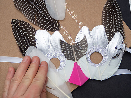 Jemand klebt eine weiße Feder von Artoz auf eine Vogelmaske aus Papier mit pinkem Schnabel. Der Untergrund ist ein braunes Kraftpapier weitere Federn sind um die Maske verteilt. Foto: Johanna Rundel