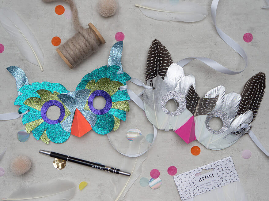 Auf dem Bild sind zwei selbstgebastelte Vogelmaske aus Papier zu sehen. Eine (links) wurde mit Halbkreisen in Grüntönen aus selbstklebendem Glitzerpapier der Marke Artoz beklebt. Der Schnabel ist orange. Die andere (rechts) wurde mit echten Federn in Naturtönen beklebt. Der Schabel ist pink. Der Untergrund ist hellgrau in Betonoptik, darauf sind buntes Konfetti und helle Pompons verteilt. Foto: Johanna Rundel