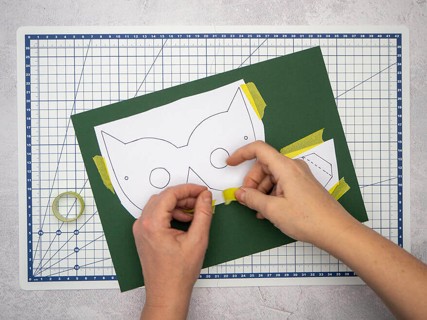 Jemand klebt mit hellgrünem Washitape eine Vorlage für eine Maske auf einen dunkelgrünen Fotokarton. Der Untergrund ist eine helle, faltbare Schneidematte mit Linien von Artoz. Foto: Johanna Rundel