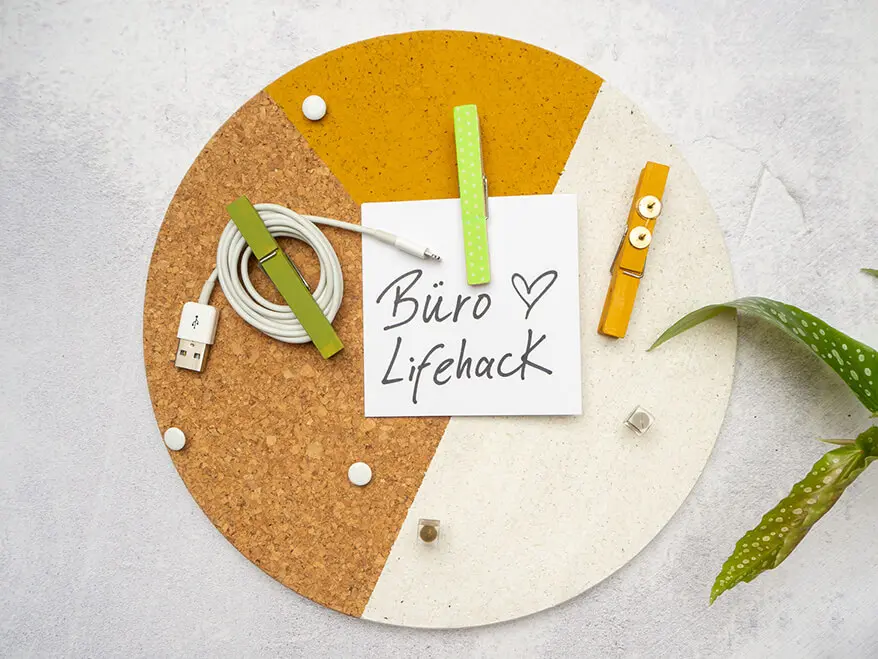 An einer grauen Wand in Betonoptik hängt eine in Weiß und Ocker bemalte Pinnwand aus Kork. Daran pinnen zwei grüne DIY-Klammerpins, die aus Wäscheklammern von Knorr Prandell gebastelt wurden. In der mittleren Klammer klemmt ein weißer, quadratischer Zettel mit der Aufschrift: Büro Lifehack. Auf die linke Klammer wurde ein weißes Kopfhörerkabel aufgewickelt. Von rechts ragt eine Zimmerpflanze ins Bild. Foto: Johanna Rundel